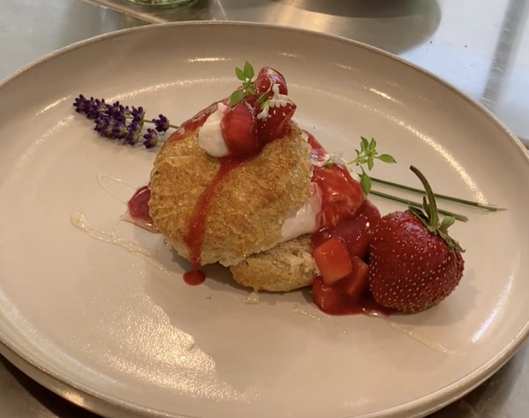 red fife wheat shortcake filled with whipped goat cheese on a plate. Drizzled with honey, strawberry mead sauce . Garnished with a fresh strawberry and two sprigs of purple edible flowers.
