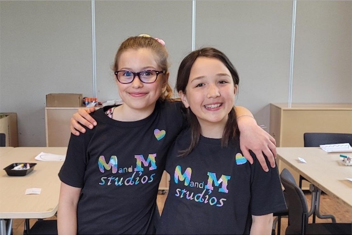 Mica and Maddie stand together smiling in black t-shirts that say "M and M Studios" in rainbow colours.